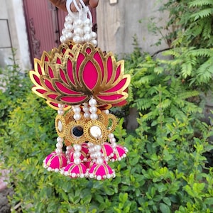 May include: A decorative garland featuring a large, pink lotus flower with gold accents. Below the lotus are gold-rimmed mirrored elements and pink bell-shaped ornaments, strung with white faux pearls. The garland is held by a white plastic fastener.