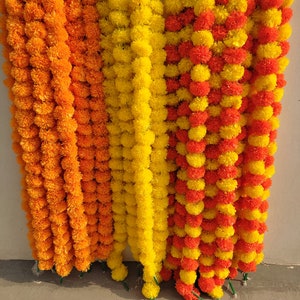 May include: A set of three garlands made of artificial marigold flowers. The garlands are in shades of orange and yellow. The garlands are hanging from a wall.