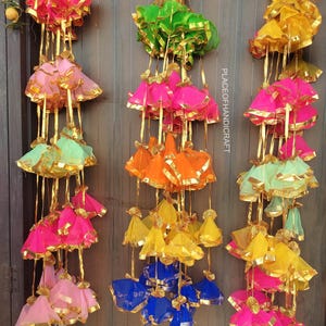 May include: A collection of vibrant, decorative hanging ornaments in various colours, including pink, green, yellow, orange, and blue. Each ornament has a layered design with gold trim and hanging gold ribbons. They are displayed against a wooden backdrop.