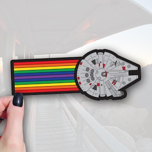 May include: A sticker featuring the Millennium Falcon spaceship with a rainbow-striped design. The spaceship is gray with red and black accents, and the rainbow stripes include red, orange, yellow, green, blue, and purple.
