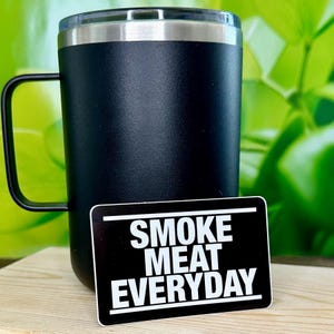 May include: Black stainless steel travel mug with a black handle. A black and white sticker with the text "SMOKE MEAT EVERYDAY" is in front of the mug.
