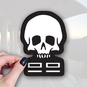May include: Black and white skull sticker with a rectangular base. The skull has a white cranium, eye sockets, and teeth, set against a black outline. The base features two horizontal white lines on a black background. The sticker is held by a hand.