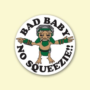 May include: A round sticker featuring a cartoon alien with green and brown skin, wearing a helmet and green armbands. The text "BAD BABY" and "NO SQUEEZIE!!" are printed around the alien in a bold, black font.
