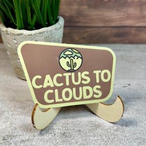 May include: A brown and light green sign with the text "CACTUS TO CLOUDS" and a cactus graphic. The sign is propped up by a small wooden stand. The background includes a small potted plant and a wooden surface.