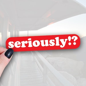 May include: A red sticker with rounded corners displays the word "seriously!?" in white, bold lettering. The sticker is held by a hand with a black painted fingernail. The background is a blurred outdoor scene.