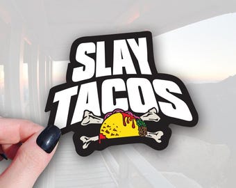 May include: A black and white sticker with the words "SLAY TACOS" in bold white letters. The sticker features a cartoon taco with a yellow tortilla, red sauce, and a pineapple. The taco is set against a background of crossed bones.