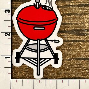 A hand holding a sticker depicting a red Weber barbecue grill with steam coming from it.