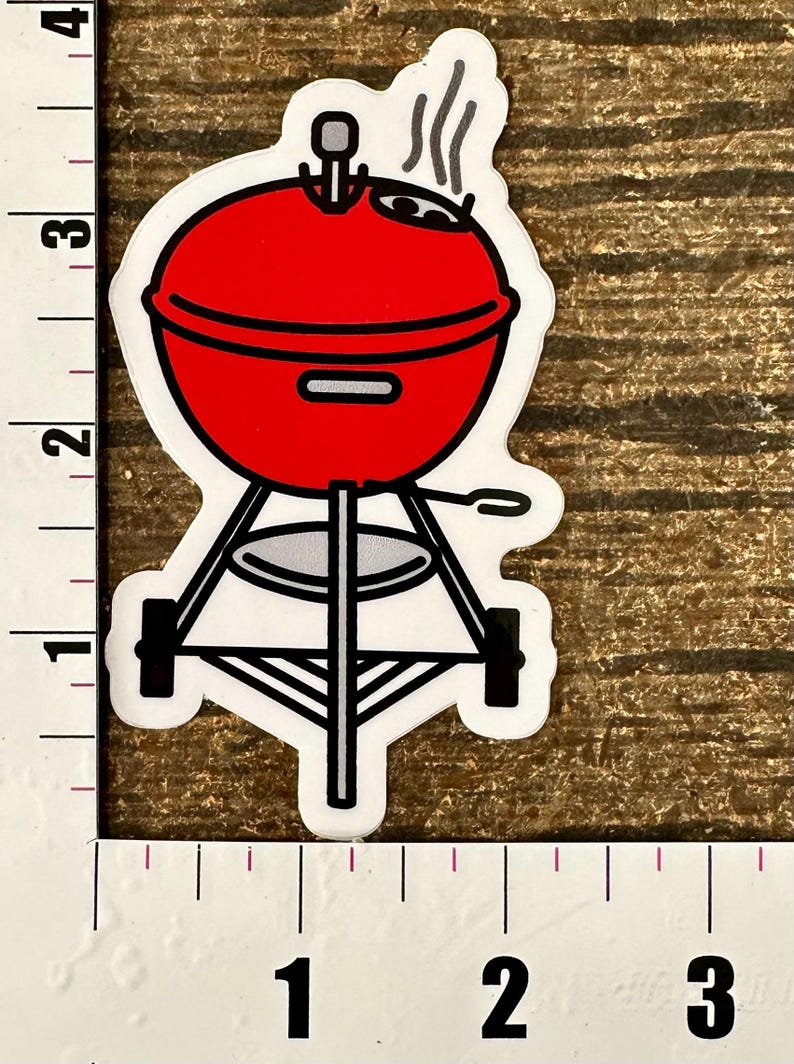 A hand holding a sticker depicting a red Weber barbecue grill with steam coming from it.