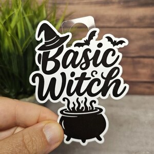 May include: A white sticker with the words "Basic Witch" in black script, featuring a witch hat, bats, and a cauldron. The sticker is held up by a clear plastic loop, perfect for hanging.