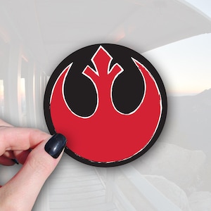 May include: A round sticker featuring the Rebel Alliance symbol from Star Wars. The symbol is red with a white outline, set against a black background. The sticker is being held by a hand with a dark nail polish.