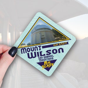 May include: A square sticker with a light blue background and a yellow border. The sticker features an image of a white observatory dome and the text "Mount Wilson, San Gabriel Mountains, Elev. 5,713 FT, Peak Slayer" in dark blue and yellow.
