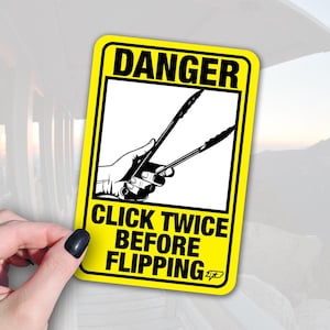 May include: A yellow sticker with black text and a black and white graphic of tongs being held by a hand. The text reads "DANGER" at the top and "CLICK TWICE BEFORE FLIPPING" at the bottom. The sticker is held by a hand with a black painted nail.