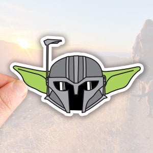 May include: A green and gray sticker of a cartoon character wearing a helmet with a visor. The character has large ears and a small mouth.