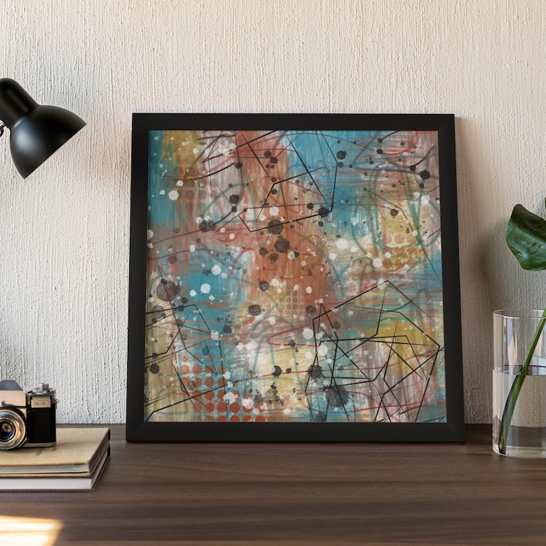 May include: Abstract art print in a black frame. The artwork features a mix of colors, including blue, orange, and white, with black lines and splatters. The painting is displayed on a wooden surface.