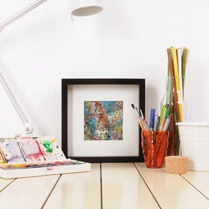 May include: A well-lit artist's workspace featuring a white desk, a white adjustable lamp, and a framed abstract painting. A watercolor palette, pens, pencils, and a small white bucket are also visible. The painting has blue, brown, and white colors.