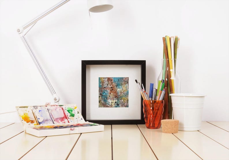 May include: A well-lit artist's workspace featuring a white desk, a white adjustable lamp, and a framed abstract painting. A watercolor palette, pens, pencils, and a small white bucket are also visible. The painting has blue, brown, and white colors.