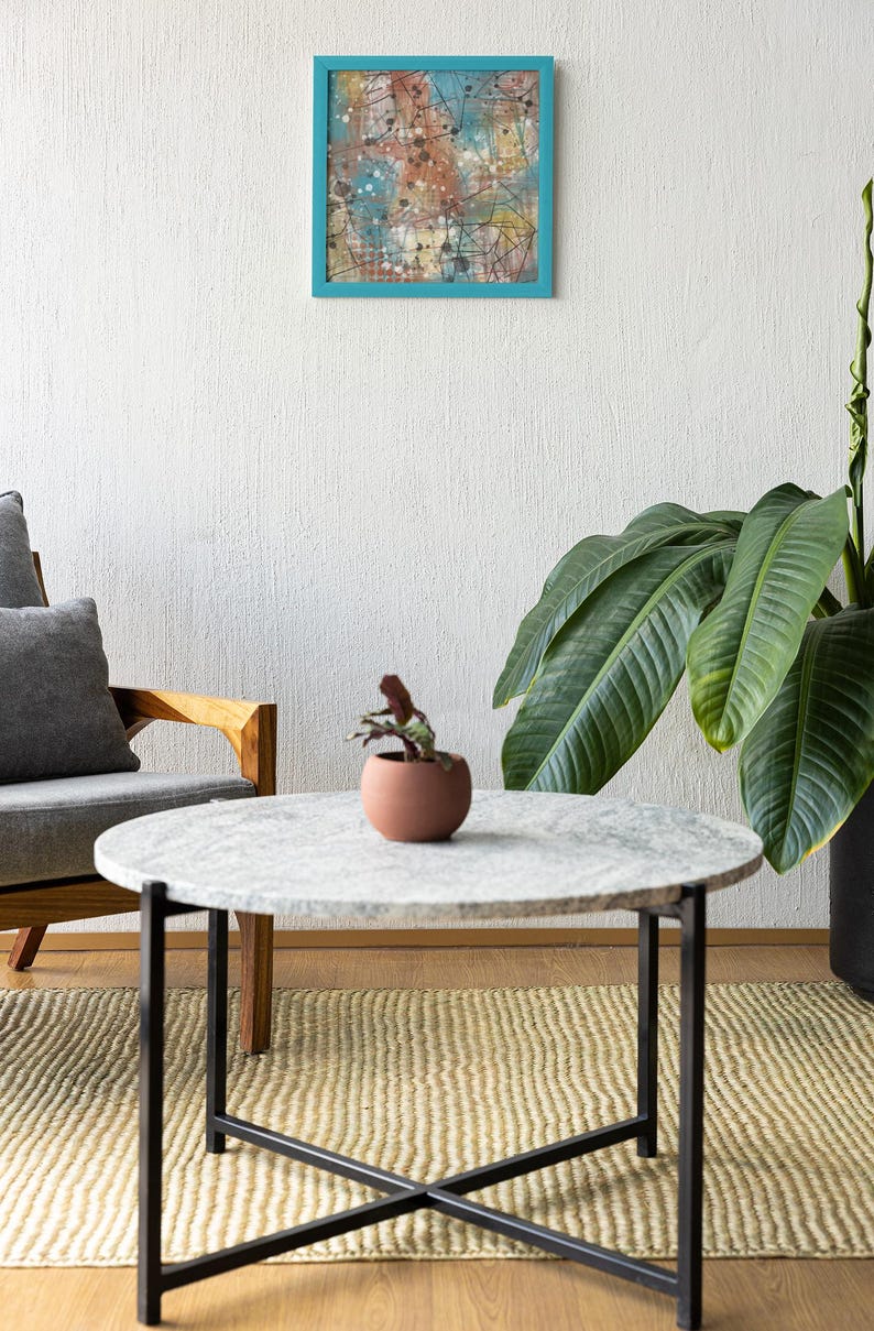 May include: An abstract painting in a teal frame hangs above a round marble-topped coffee table with black metal legs. A small potted plant sits on the table. A gray armchair and a large potted plant are also in the room.