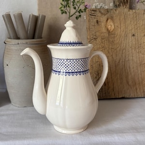 Puede incluir: Una cafetera de cerámica blanca con un patrón geométrico azul alrededor del centro y en la tapa. La cafetera tiene un pico curvo y un asa. Otros elementos en la imagen incluyen una tabla de cortar de madera, un jarrón de cerámica con velas y una botella de cerámica alta.