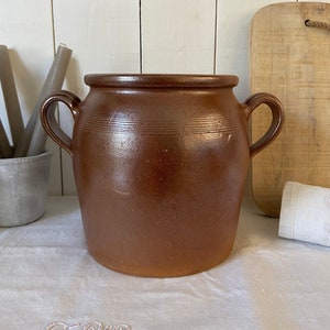 Puede incluir: Una gran olla de barro marrón con dos asas. La olla tiene una forma ligeramente redondeada y está hecha de arcilla rugosa y texturizada.