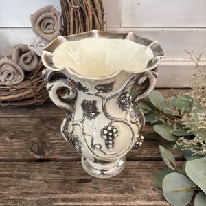 Peut inclure: Un vase en céramique de couleur crème avec un bord ondulé et deux anses latérales. L'extérieur présente des décorations en relief de couleur sombre représentant des grappes de raisin et des feuilles de vigne sur un pied circulaire.