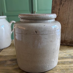 Puede incluir: Jarra de gres antigua de color beige, con cuerpo redondeado y borde ligeramente ensanchado. La jarra está marcada con el número "79". Esta antigua jarra de almacenamiento tiene una forma cilíndrica simple y una estética rústica.