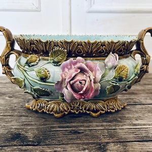 Peut inclure: Un centre de table en céramique avec un intérieur et un extérieur bleu clair, orné d'une grande rose rose et de feuilles vertes. Le bol a des poignées et des bordures marron, posé sur une surface en bois.