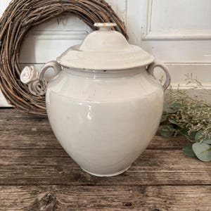 Op de afbeelding: Een crèmekleurige keramische pot met deksel, een ronde vorm, twee handvatten aan de zijkanten en een bijpassend deksel met een kleine knop. Het oppervlak vertoont zichtbare fijne haarscheurtjes en lichte gebruikssporen langs de rand.