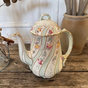 Peut inclure: Une théière vintage en céramique avec un couvercle. La théière présente un motif à rayures bleu clair et blanc avec des accents floraux roses, jaunes et verts. Des bordures dorées ornent le bec, la poignée et le couvercle. La théière est sur une surface en bois.