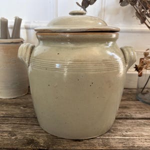 May include: A large, antique stoneware crock with a lid, featuring a light beige glaze and horizontal banded detailing. The crock has two side handles and a matching lid. A smaller, similar jar is visible to the left, along with a vase of dried flowers.