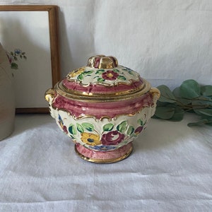 Peut inclure: Un plat couvert en céramique vintage avec une base rose et un motif floral blanc. Le plat est orné d'accents dorés et d'un couvercle avec un motif floral.