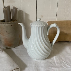 Peut inclure: Une cafetière en porcelaine blanche avec un motif en spirale. La cafetière a un couvercle et une poignée incurvée. Un petit pot en céramique grise avec des bougies grises est en arrière-plan. Un tissu blanc plié est au premier plan.