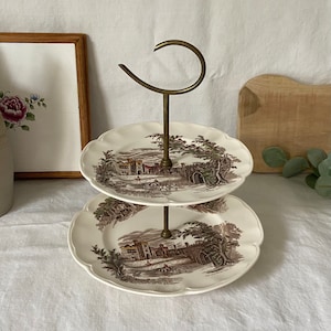 Puede incluir: Un soporte para tartas de dos niveles con un asa de latón. El soporte está decorado con un patrón floral marrón y blanco y presenta una escena de un castillo.