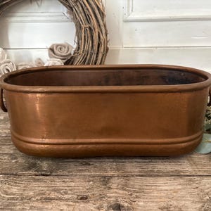Pode incluir: Uma bacia oval de cobre com um acabamento metálico liso e um tom castanho-avermelhado quente. O recipiente inclui um rebordo reforçado, uma faixa horizontal em relevo perto da base e pequenas pegas de metal fixadas em cada lado do objeto.