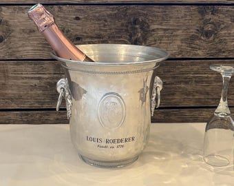 May include: A silver champagne bucket with a bottle of champagne inside. The bucket has handles and the words "LOUIS ROEDERER Fondé en 1776" are printed on the front. A wine glass is next to the bucket.