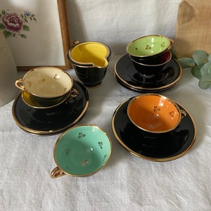 Puede incluir: Un juego de cinco tazas de té negras y platillos con ribete dorado. Cada taza tiene un interior de color diferente: crema, verde, naranja y azul. Las tazas tienen un patrón floral dorado.