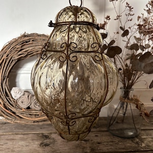 May include: A vintage-style glass lantern with a decorative metal frame. The lantern has a bulbous shape with a textured surface and is suspended by a metal ring. The glass has a warm, amber hue, and the metal frame features ornate scrollwork.
