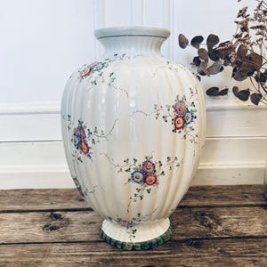 Peut inclure: Un grand vase en céramique blanc cassé avec un corps bulbeux et un design cannelé. Le vase est orné de motifs floraux peints à la main dans des tons de rouge, bleu et violet, avec des vignes vertes. La base et le bord ont des accents verts. Un objet de décoration.