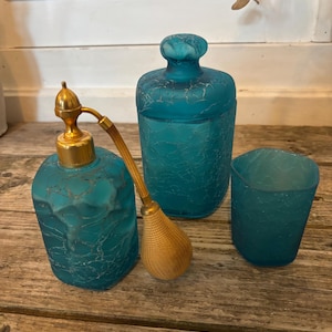 Peut inclure: Un ensemble de flacons de parfum en verre bleu sarcelle antique et un petit gobelet. Le flacon de parfum de gauche a un atomiseur doré. Le verre a un effet craquelé. L'ensemble est présenté sur une surface en bois.