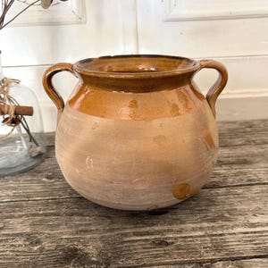 Puede incluir: Una rústica olla de barro con dos asas y cuerpo redondeado. La olla presenta una base marrón claro con un borde y asas de color marrón más oscuro. Se encuentra sobre una superficie de madera envejecida, lo que sugiere una estética vintage o antigua.