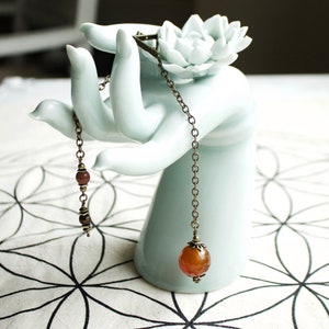 May include: A delicate necklace with a carnelian gemstone pendant hangs from a ceramic hand sculpture. The necklace features a chain with small brown beads and a bronze clasp.