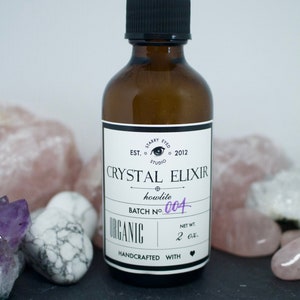 May include: A brown glass bottle with a dropper top and a white label that reads "Crystal Elixir howlite Batch No. 004 Organic Handcrafted with" and a heart symbol. The bottle is sitting on a black surface with various crystals.