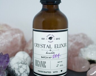 May include: A brown glass bottle with a dropper top and a white label that reads "Crystal Elixir howlite Batch No. 004 Organic Handcrafted with" and a heart symbol. The bottle is sitting on a black surface with various crystals.