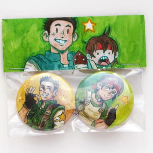 May include: Two round, colorful pinback buttons featuring cartoon illustrations of characters. The buttons are yellow and green, with detailed drawings of the characters. The packaging includes a green background with additional character illustrations.