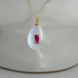 May include: A gold chain necklace with a pear-shaped pendant. The pendant is white with a rectangular, magenta-colored stone set in the center. The necklace is displayed against a white background.