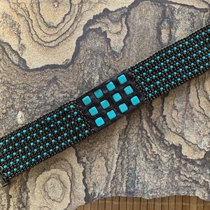 May include: A black beaded bracelet with a turquoise square design. The bracelet has metal clasps on each end.