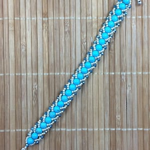 May include: A turquoise and silver beaded bracelet with a toggle clasp. The bracelet is made with small round beads and is on a bamboo background.
