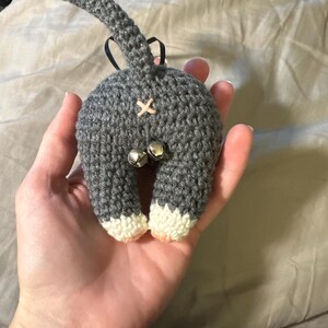 May include: A gray crocheted cat butt ornament with a tail, two bells, and white-tipped feet. The ornament has a pink cross stitch detail and a black ribbon for hanging. A unique, handmade cat-themed decoration.
