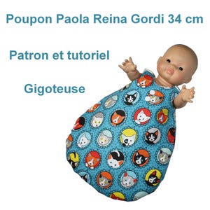 May include: A Paola Reina Gordi doll, 34 cm tall, wearing a blue cat-themed sleep sack. The sleep sack features a repeating pattern of cat faces in various colors. Text on the image reads "Patron et tutoriel" and "Gigoteuse".