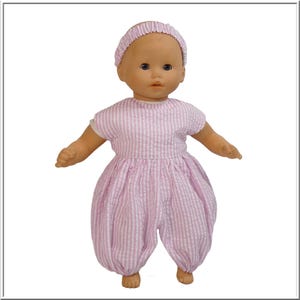 Könnte beinhalten: Eine Puppe in einem rosa-weiß gestreiften Strampler und einem passenden Stirnband. Die Puppe hat blaue Augen und einen neutralen Gesichtsausdruck. Das Outfit ist einteilig mit kurzen Ärmeln und einem ballonartigen Unterteil.