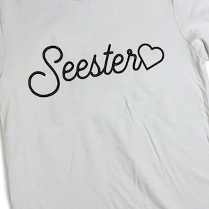 以下が含まれることがあります： 白いTシャツに、黒い筆記体で「Seester」と書いてあり、「r」の後に小さなハートマークがついています。